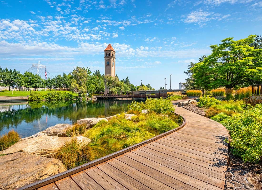 About Our Agency - View of a Wooden Trail Surrounded by Green Folaige Next to a Pond in a Park on a Sunny Day with the Spokane Clock Tower Visible in the Distance